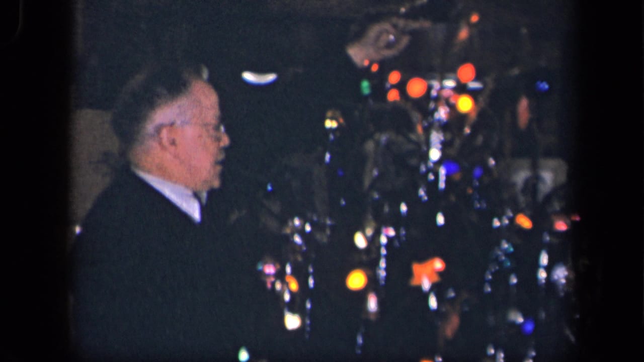 The older man remains beside the illuminated Christmas tree, with colored bulbs and shiny decorations visible in the dim home interior.