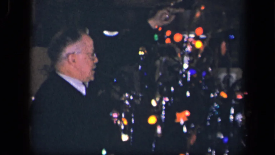 The older man remains beside the illuminated Christmas tree, with colored bulbs and shiny decorations visible in the dim home interior.