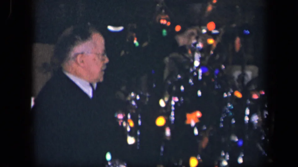 An older man in glasses is seen in profile near a decorated indoor Christmas tree, with tinsel and multicolored lights catching the film exposure.