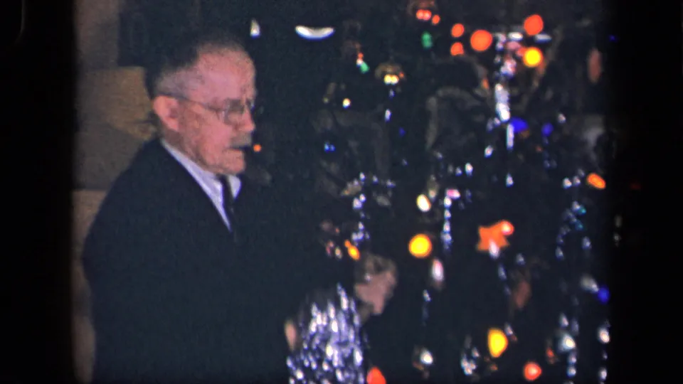 The older man turns more toward the camera while standing beside the Christmas tree, where tinsel, ornaments, and colored lights fill the frame.