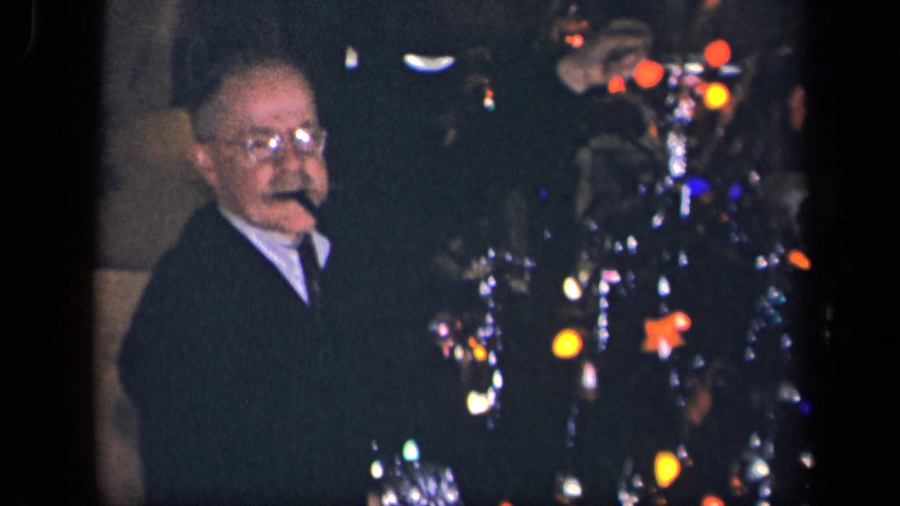 Man with glasses faces the camera near a brightly lit Christmas tree.