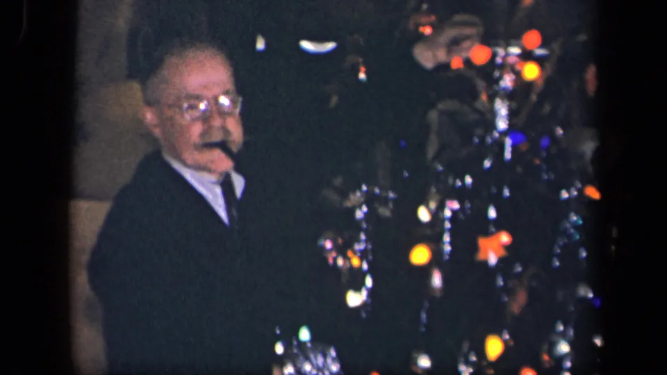 The man faces toward the camera beside the decorated Christmas tree, with colored lights and ornaments visible in the dim 1942 interior.