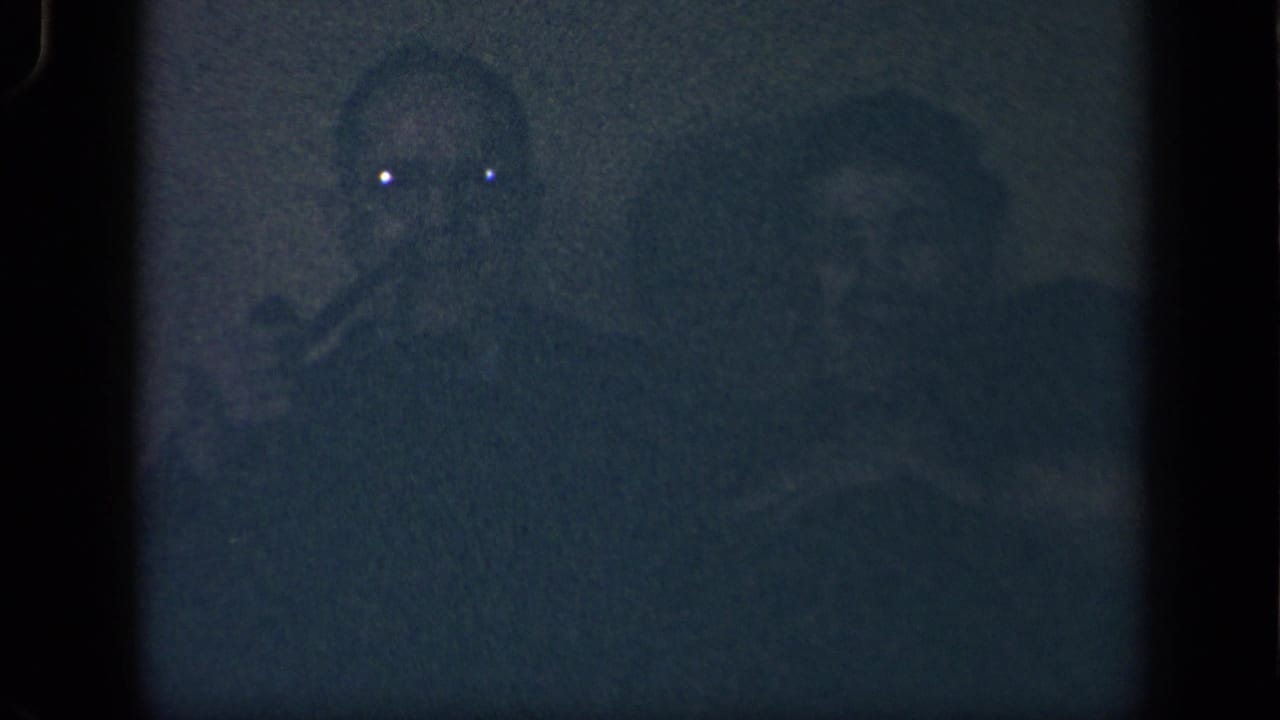 A man with a pipe and a seated woman appear in a grainy, dark blue 8mm frame.
