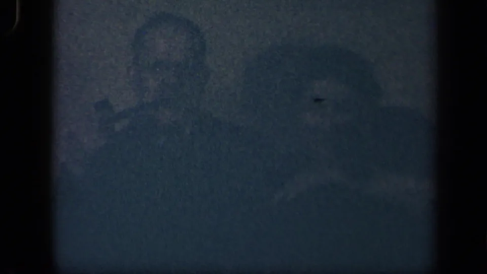 Dark 8mm frame from the Sebring, Ohio clip showing the seated man in shadow and another adult figure faintly beside him.