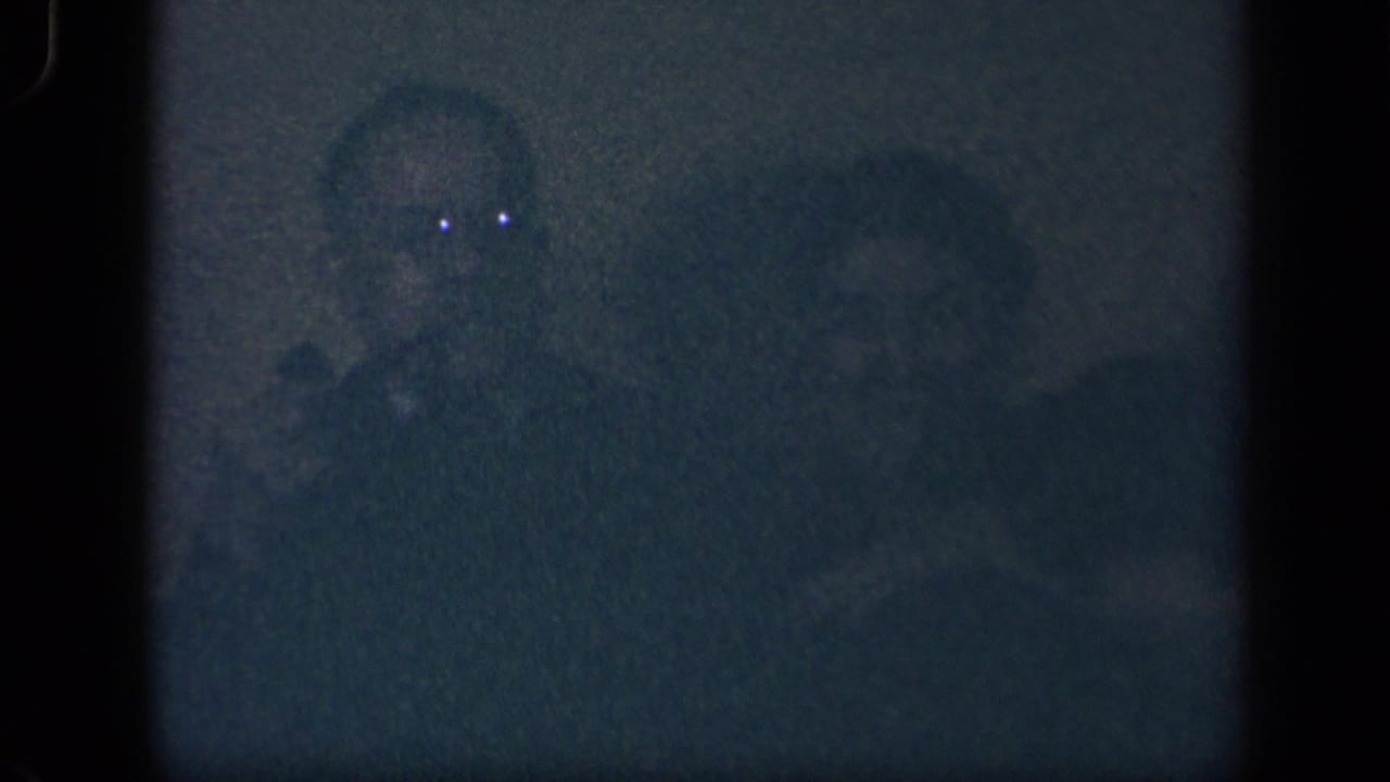 Shadowed adults sit in a dim room, with one face faintly visible.