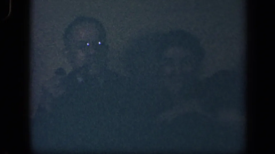 The dim 8mm frame shows adults seated in a private interior, with facial details softened by grain, darkness, and film exposure.