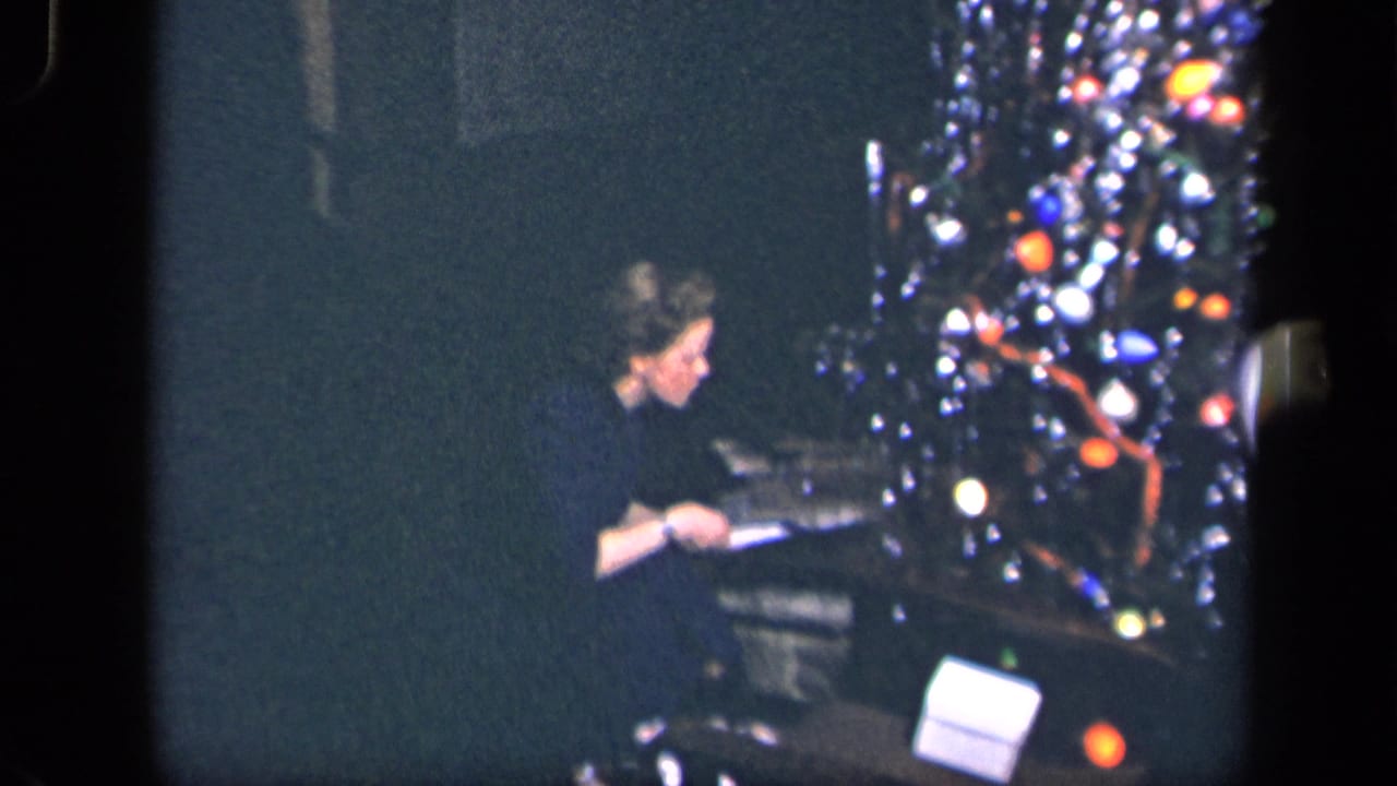 Woman seated beside a lit Christmas tree while handling a gift in a dim room.