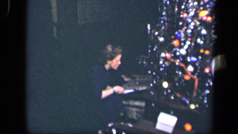 In 1942 Sebring, Ohio home-movie footage, a woman in a dark dress sits near a lit Christmas tree and begins opening a present.