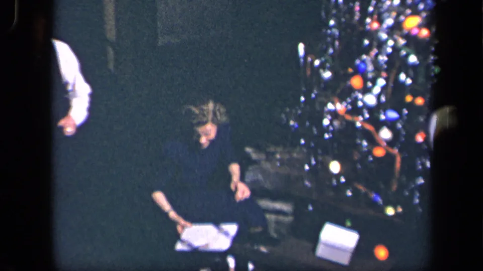 The seated woman leans over wrapping paper near the illuminated Christmas tree in the Sebring, Ohio holiday home movie.