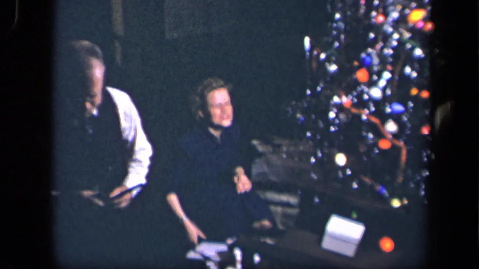 A man in a white shirt and dark vest stands close to the seated woman as she smiles beside the decorated Christmas tree.