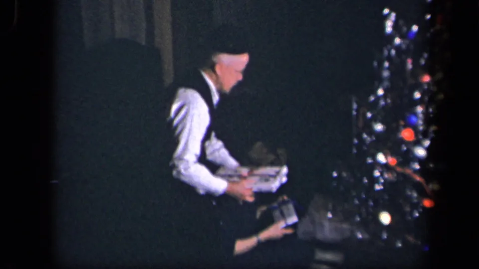 The man holds the wrapped gift close to the seated woman as the decorated Christmas tree fills the side of the frame.