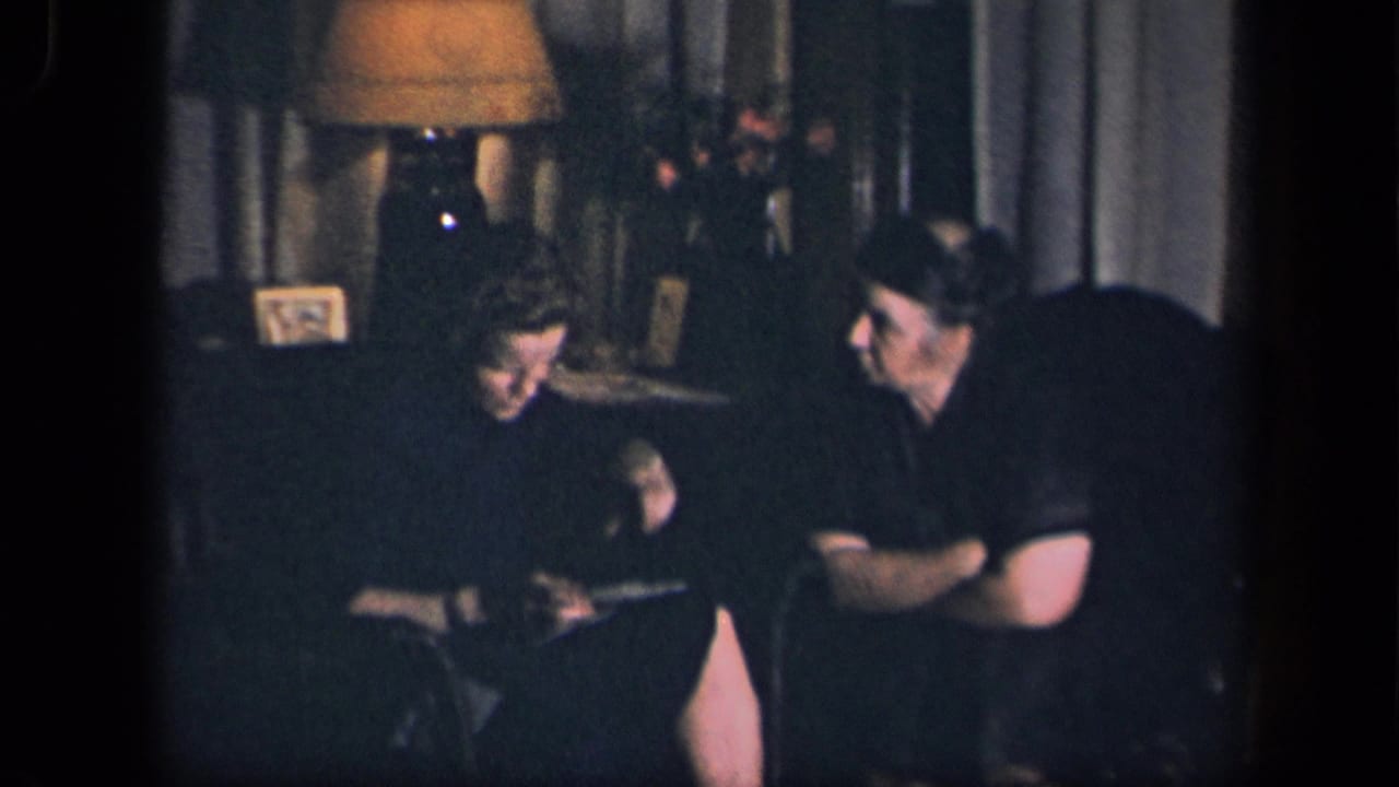 Two adults sit in a dark living room near a lamp while the woman looks down at something in her hands.
