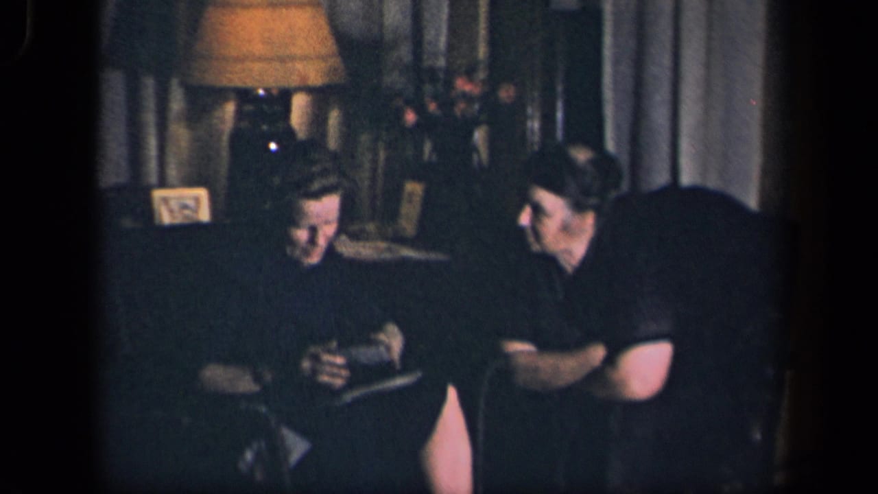 A woman holds a small object while a man seated beside her watches in a dimly lit room.