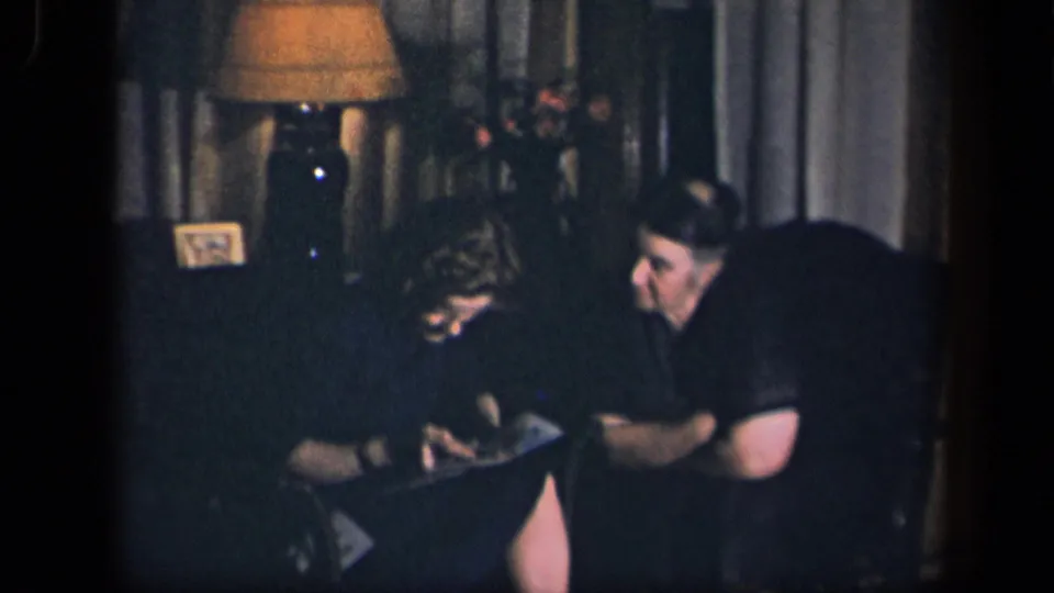 In the low-lit 1942 home interior, the woman bends forward over an item on her lap while the man watches from the side.