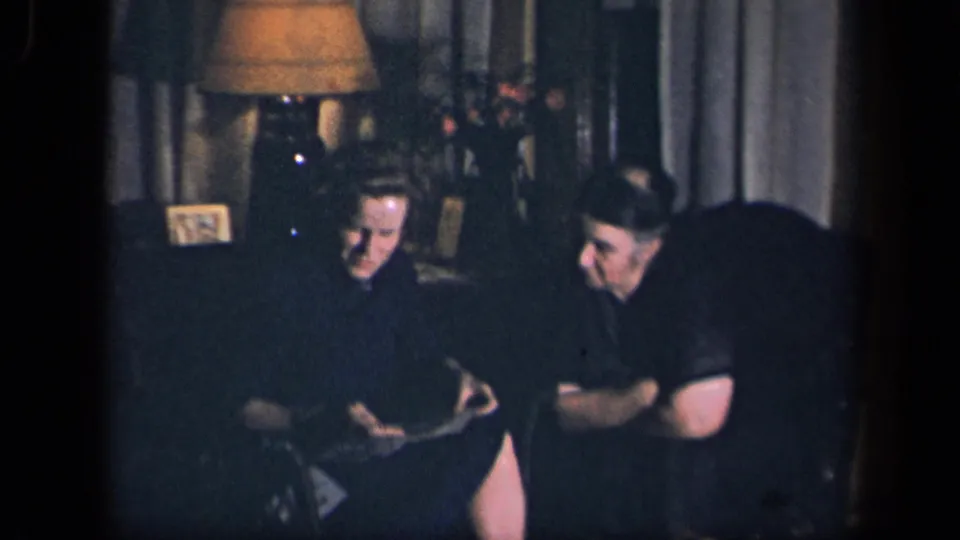 In this 1942 Sebring, Ohio home-movie frame, a woman in a dark dress sits beside a man while looking down at a card in a dim living room.