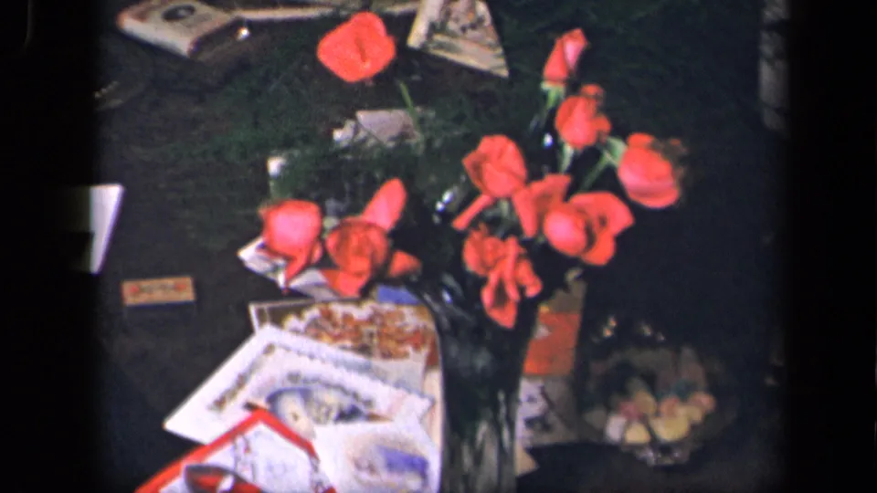 A close view of a living-room table shows red roses in a vase, cards or papers, and small household objects in the same 1942 home-movie scene.