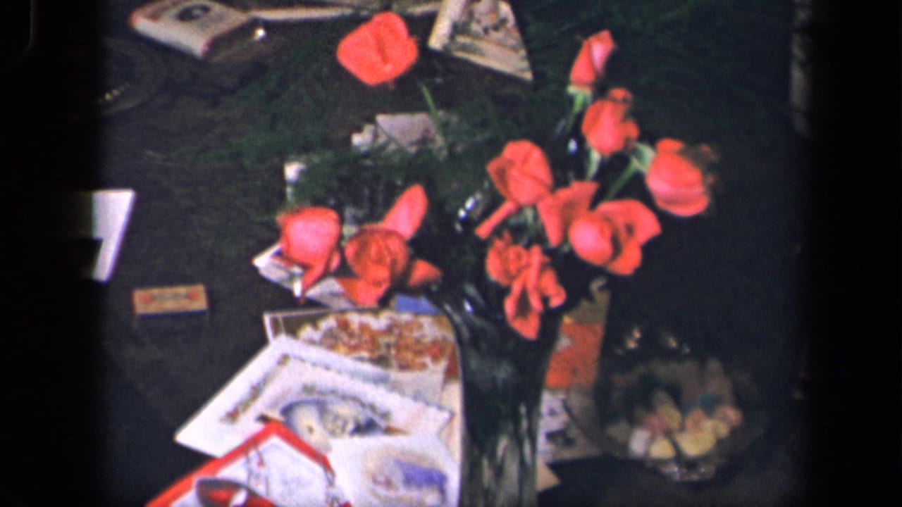 1942 Sebring, Ohio 8mm frame showing red roses in a vase surrounded by greeting cards, printed papers, and a candy dish on a dark tabletop.
