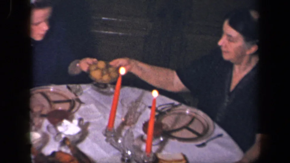Two women are shown mid-exchange at a dim dinner table with red candles, plates, glassware, and a white cloth.