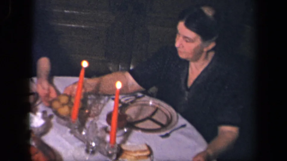 A woman reaches near a dish on the candlelit table, with another person partly visible at the edge of the frame.