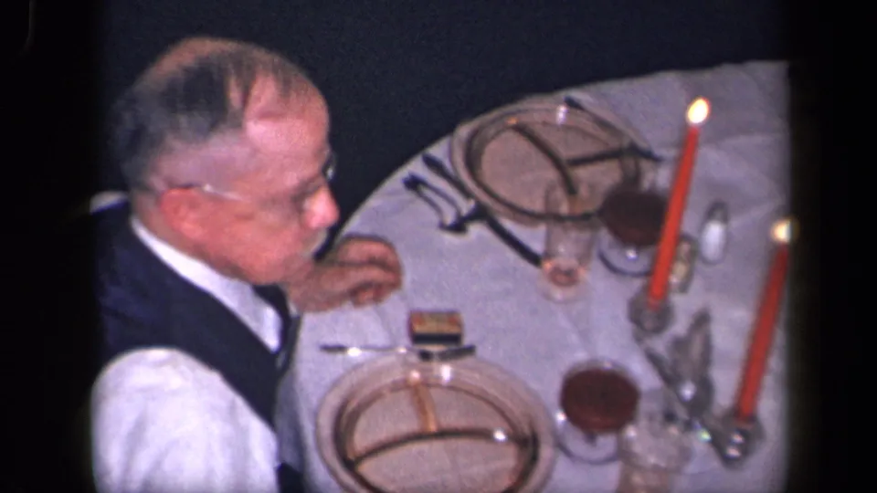 An older man wearing glasses and a vest sits at a candlelit dinner table with divided plates, glasses, and a white tablecloth.