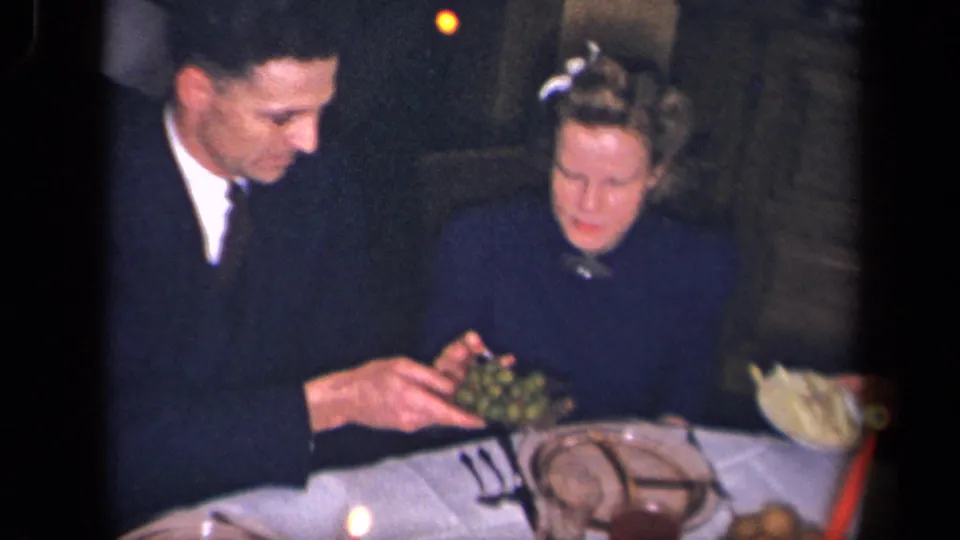 In the Sebring, Ohio dinner scene, a suited man passes a dish of olives to a woman near plates, cutlery, and candlelight.