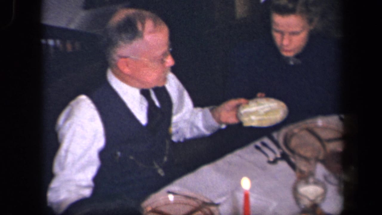 An older man in a white shirt and dark vest serves food at a candlelit family dinner table in the Sebring, Ohio home-movie footage.