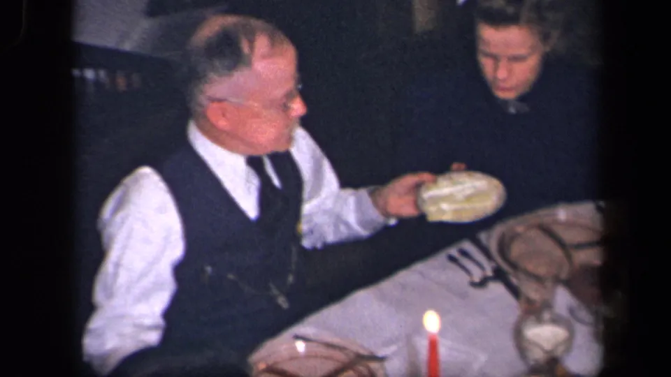 An older man in a white shirt and dark vest serves food at a candlelit family dinner table in the Sebring, Ohio home-movie footage.