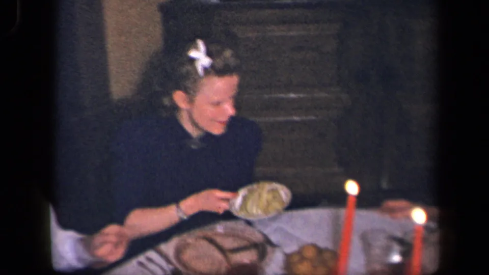 A woman with a white flower in her hair sits at the family dinner table, with plates, candles, and serving dishes visible.