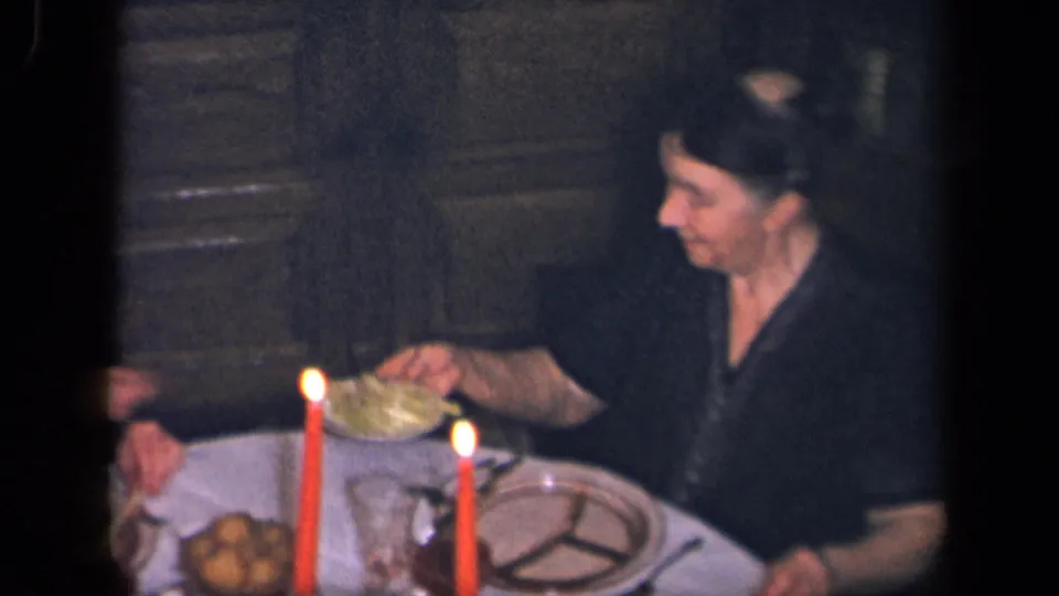 A woman in a dark dress sits at the candlelit family meal, with red candles, plates, and a serving dish on the table.