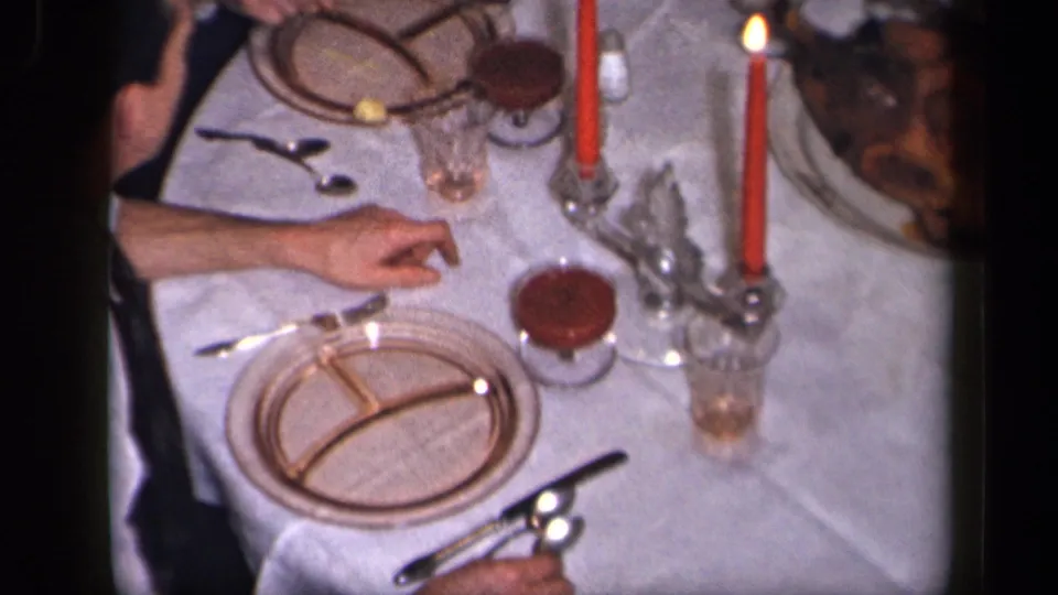 The frame shows a candlelit dinner table in 1942 Sebring, Ohio, with divided plates, glassware, food, and a person's arm near the setting.
