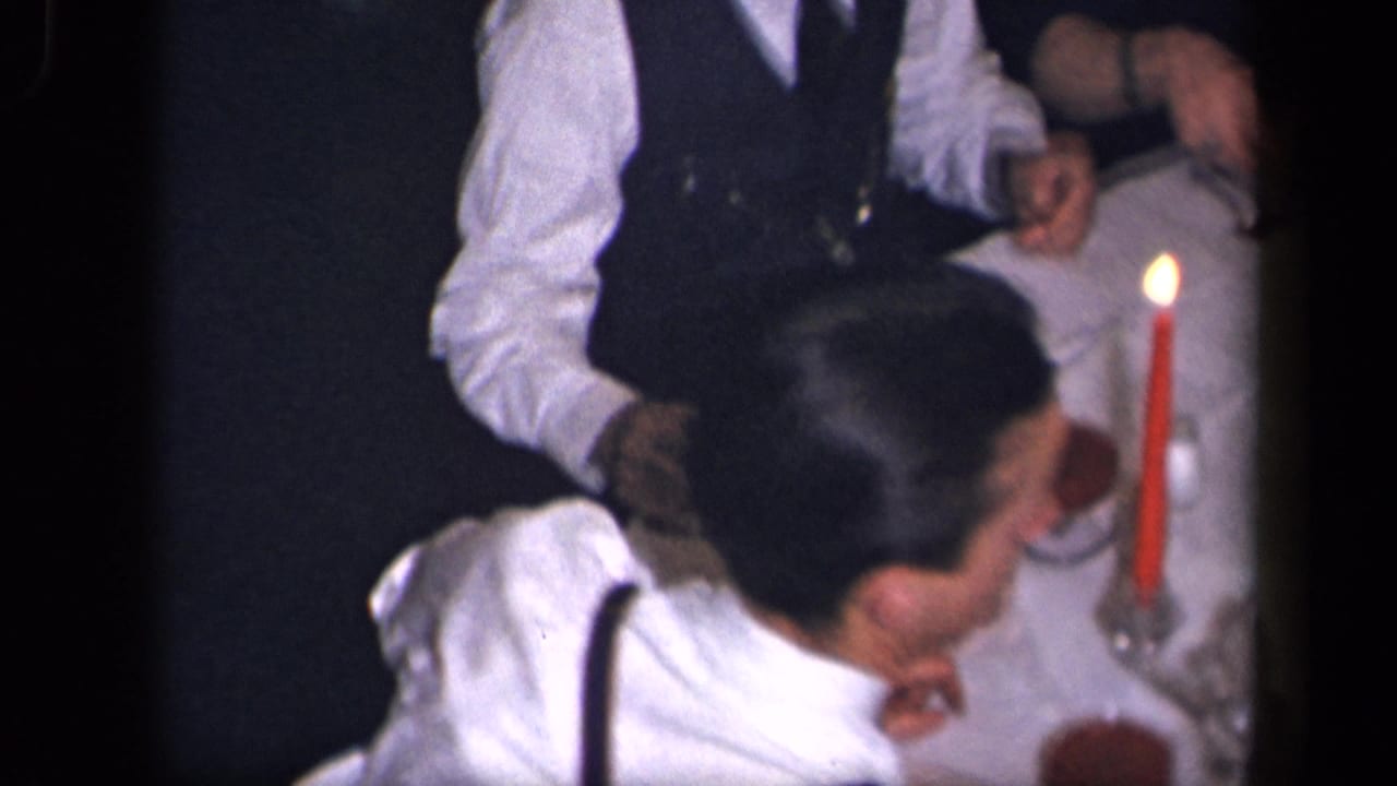 Man in white shirt and suspenders seated near a candlelit dinner table.