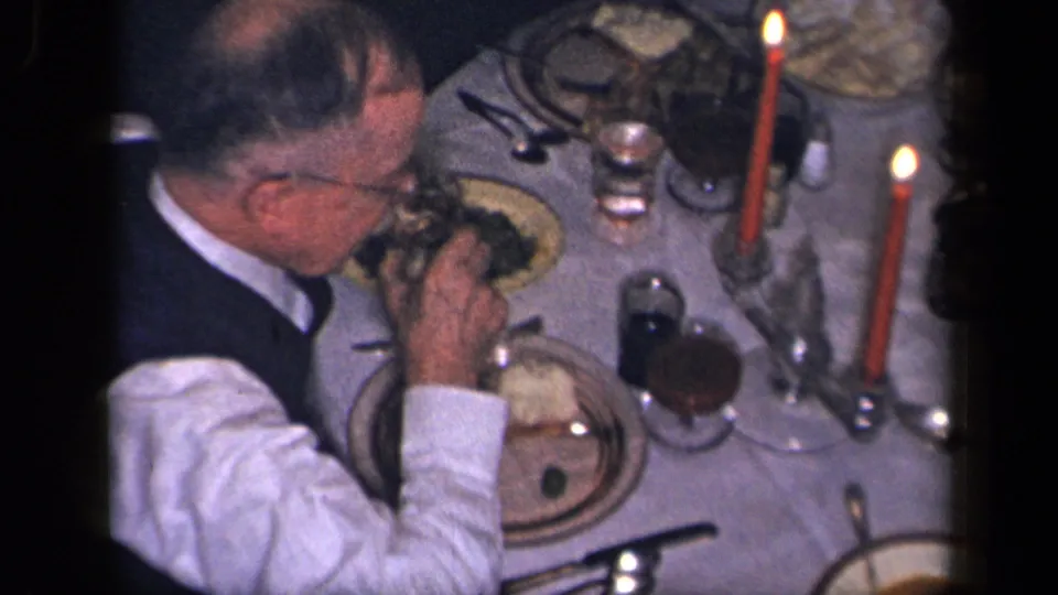 Overhead 8mm frame of a man eating at a candlelit dining table with plates, glasses, serving dishes, and a tablecloth.