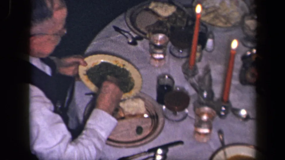 Candlelit home-movie frame showing a man reaching toward his plate at a formal family dinner table in Sebring, Ohio, 1942.