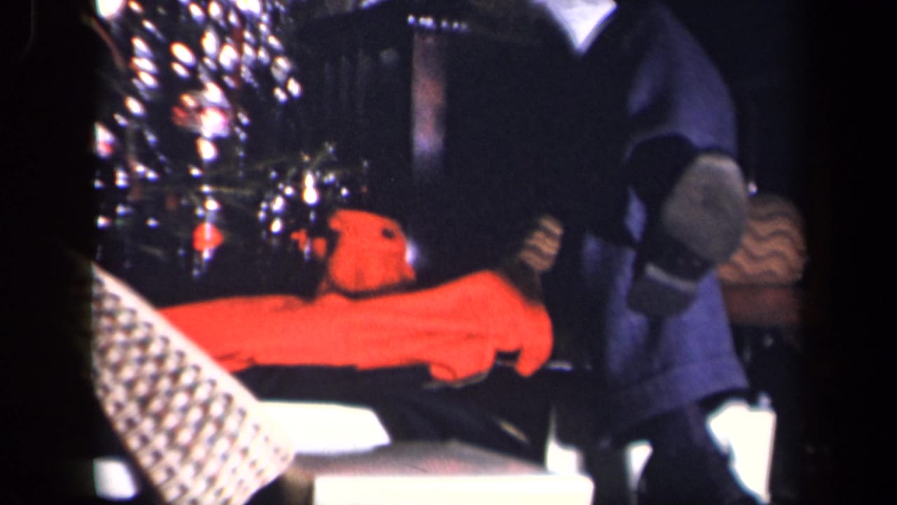 A closer home-movie view shows the decorated tree, a bright orange plush or toy-like object on furniture, and the seated man's legs at the edge of the frame.
