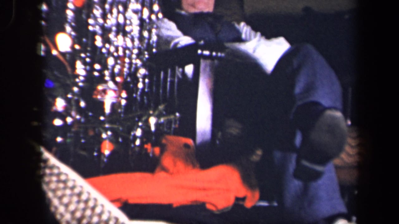The 8mm frame catches part of the seated man, the tinsel-covered Christmas tree, and a bright orange plush or toy-like object in the private living room.