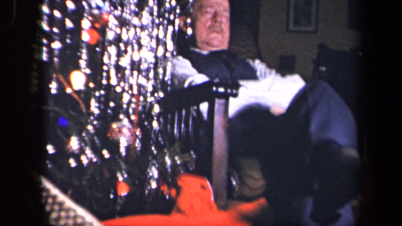 An older man reclines in a dark chair near the lit and tinsel-covered Christmas tree, with the orange toy-like object visible low in the frame.