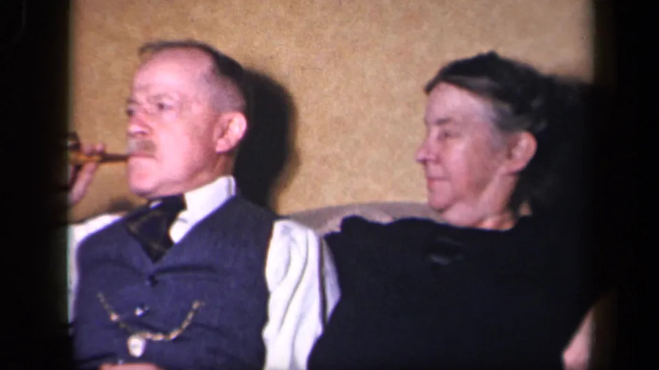 The older man smokes a cigar while seated beside an older woman in a 1942 Sebring, Ohio home interior.