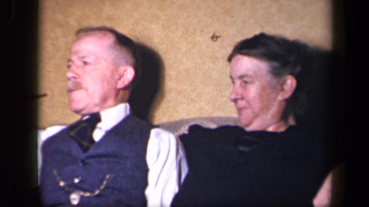 In 1942 Sebring, Ohio home-movie footage, an older couple sits closely indoors as the man in a dark vest and tie looks left.