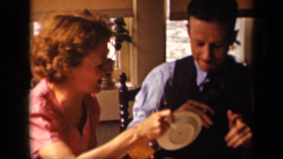 The woman smiles across the table while the man in a vest looks down at a small plate inside the 1942 Sebring home-movie setting.