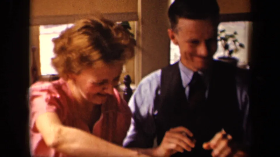 The woman leans forward across the breakfast table as the man smiles, captured in warm 8mm kitchen light.