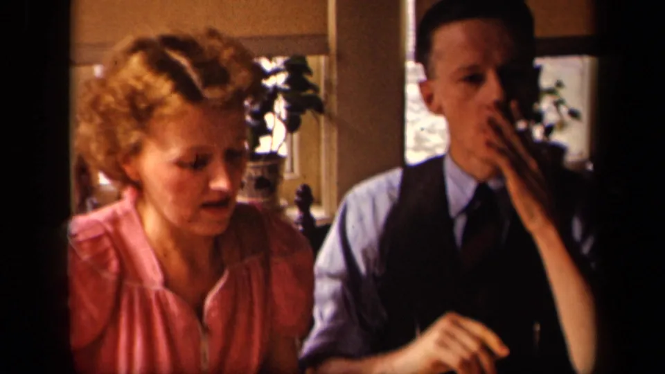 At the kitchen table, the man raises a cigarette to his mouth while the woman looks down near the breakfast setting.