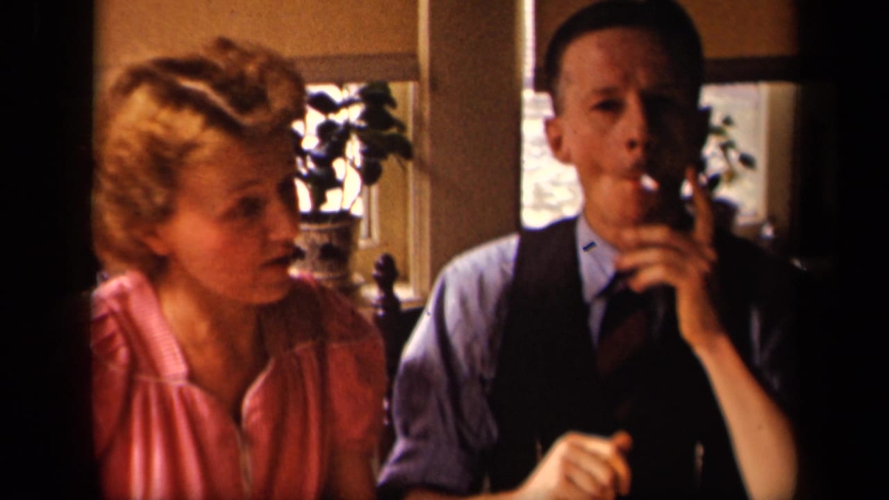 Woman in a pink blouse sits beside a man in a vest who smokes at a table.