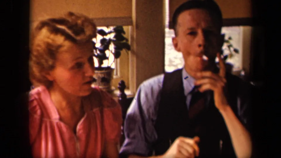 In a 1942 Sebring, Ohio home interior, a woman in pink sits close to a man in a vest as he raises a cigarette.