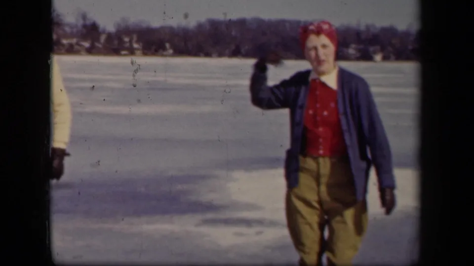 A woman in a red headscarf and blue cardigan gestures on the frozen lake, with a man partly visible at the frame edge.