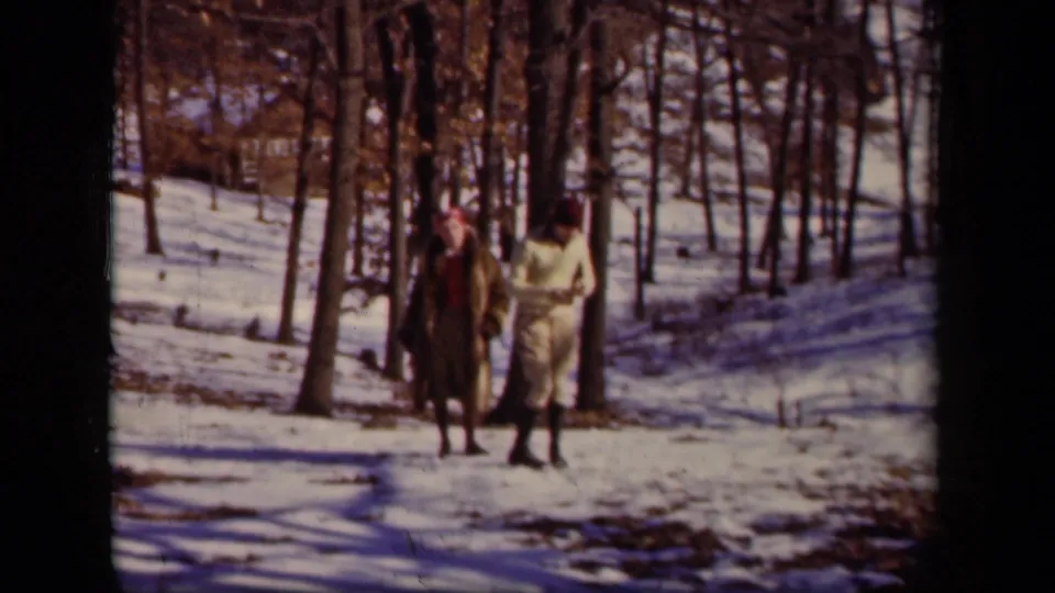 The couple moves through a snow-covered wooded area in the 1942 Ohio home-movie footage, framed by bare tree trunks and winter light.
