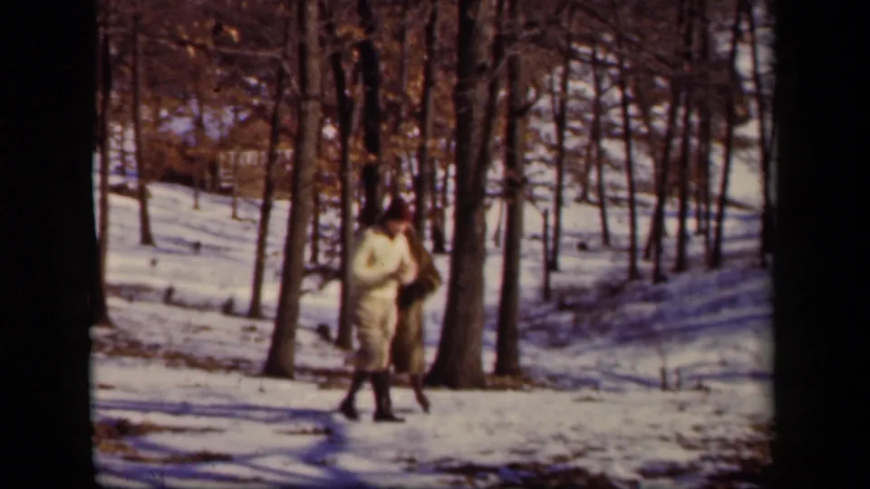 A woman in winter clothing walks through a snowy forested area, with bare trunks and patches of sunlight visible around her.