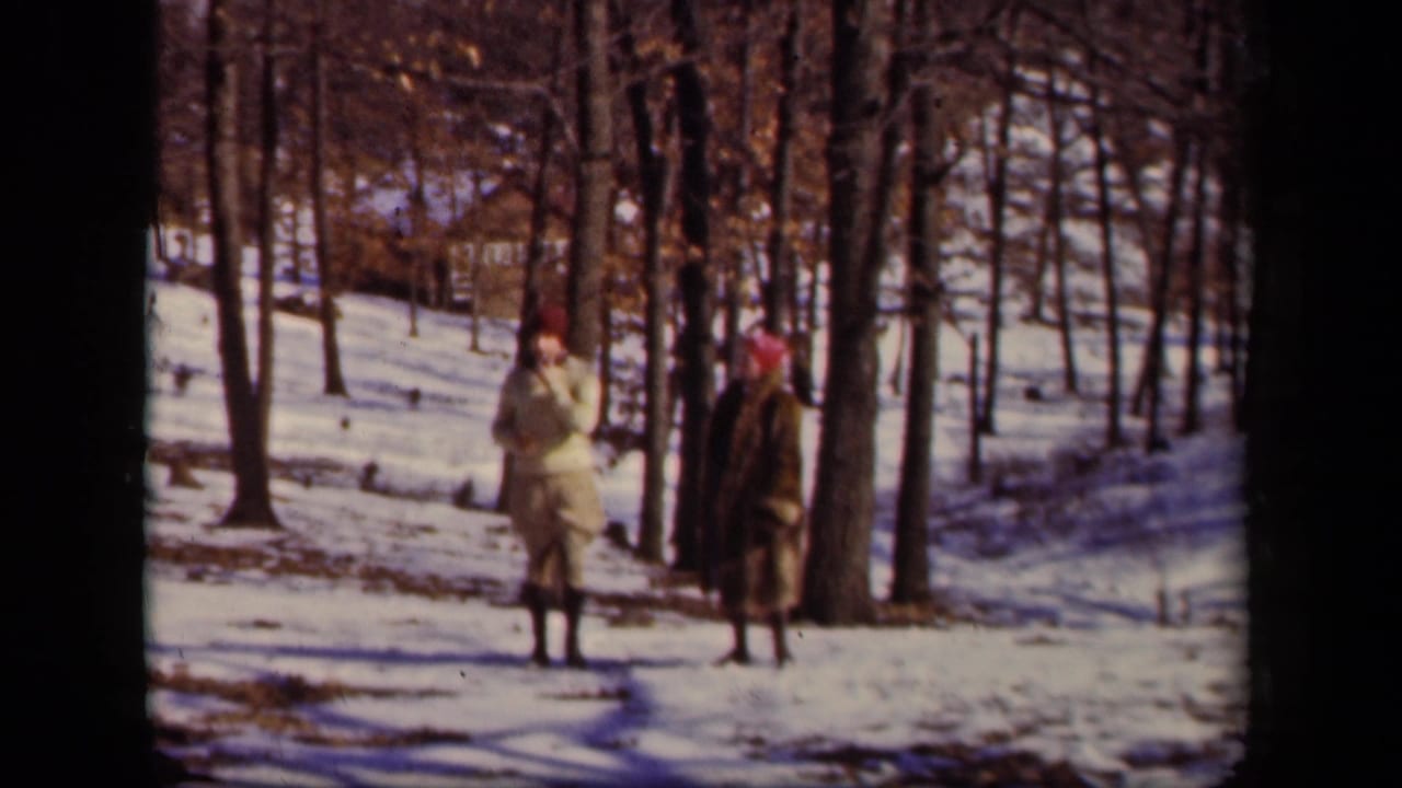 Two adults walk through a snowy forest with bare trees behind them.