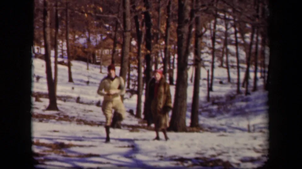 A man in a light coat and a woman in a darker coat walk together through snowy woods in the 1942 home-movie footage.