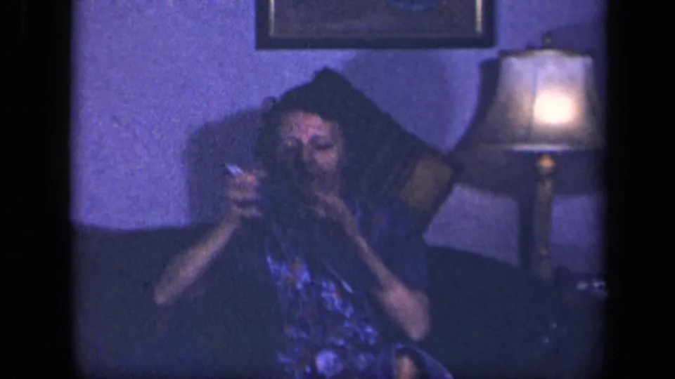 The woman appears to inspect or work on her knitting while seated in a Sebring, Ohio living room, with a lamp glowing beside her.