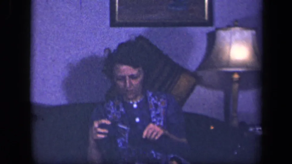 A 1942 home-movie frame shows the woman seated beneath a framed picture, working with a knitting project in a blue-tinted living room.